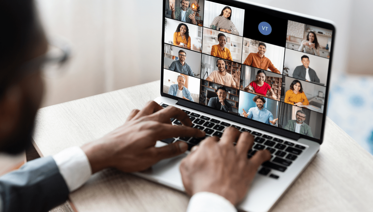 table in front of an open laptop, actively participating in a video conference. The laptop screen displays a grid of various participants, each in their own frame